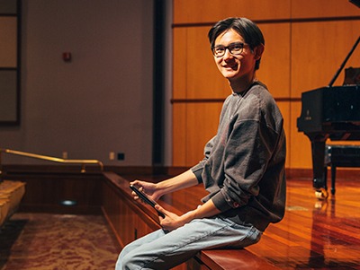 Male student sitting on the edge of a stage with a grand piano in the background