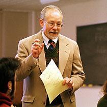 A person speaking in a classroom holding papers.