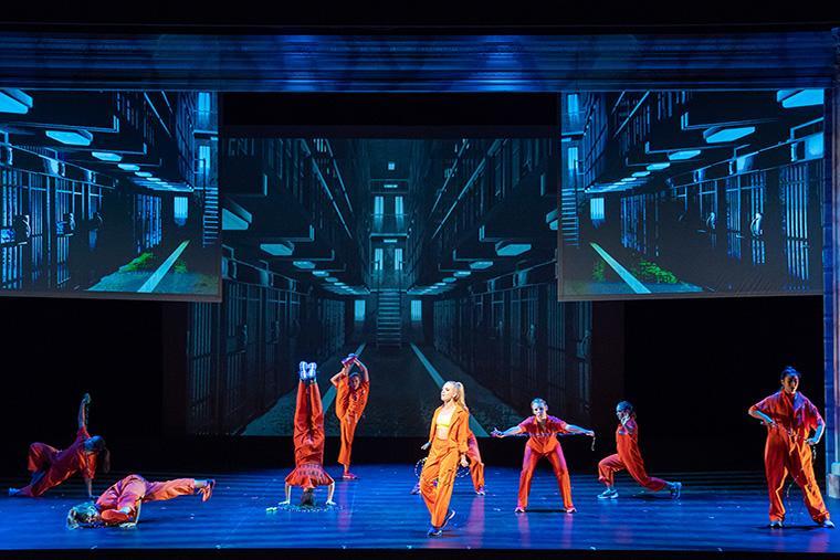 Stage performance of Legally Blonde with actors in pink-orange outfits against a blue backdrop.