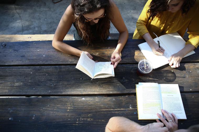 Students discussing notes while seated at a wooden table outdoors.