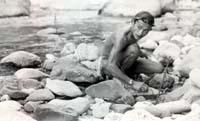 A person sitting by a stream with rocks, touching the water.