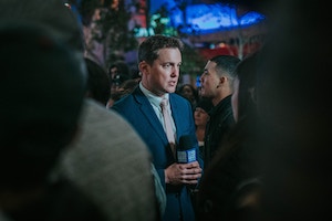 Male journalist in a suit and tie, holding a microphone while he speaks 