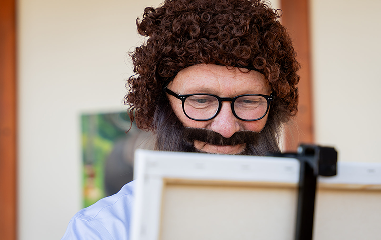 A man in a curly brown, Bob Ross-esque wig and fake brown wig focuses on a canvas.