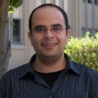 A person wearing glasses, standing outdoors, with greenery and a building in the background.