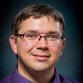 A person wearing glasses and a purple shirt, against a dark background.